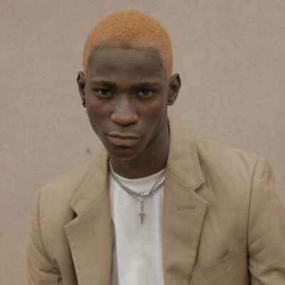 Confident African American male with dyed hair wearing trendy suit and chain looking at camera while standing near wall on street