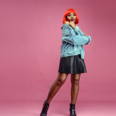 Full body of young African American female with dyed hair wearing denim jacket and mesh tights looking at camera on pink background in studio