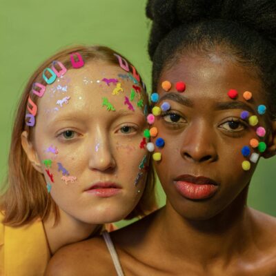 Trendy African American woman with bright pom pom on face standing near female model with hairpins and colorful stickers looking at camera against green background